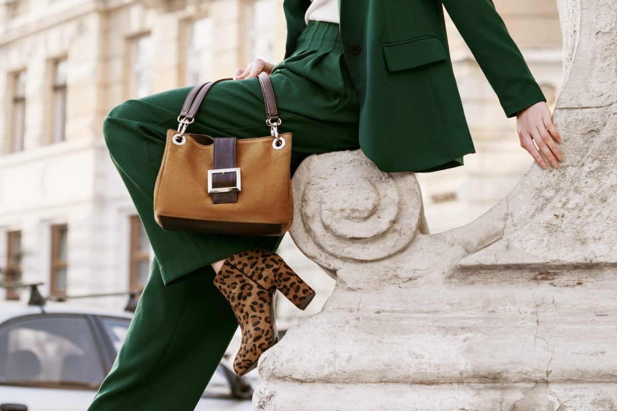 Outdoor full body fashion portrait of  fashionable woman wearing sunglasses, white turtleneck, green suit, blazer, trousers, leopard print boots, holding suede bag, posing in street of european city